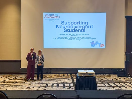 Two women standing smiling in front of a large projection screen reading "Supporting Neurodivergent Students: Lessons Learned from Year 1 of the SHINE Program" at a conference.