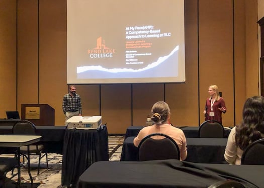 A man and a woman presenting in a conference room with a projection screen that reads "At My Pace (AMP): A Competency-Based Approach to Learning at RLC."