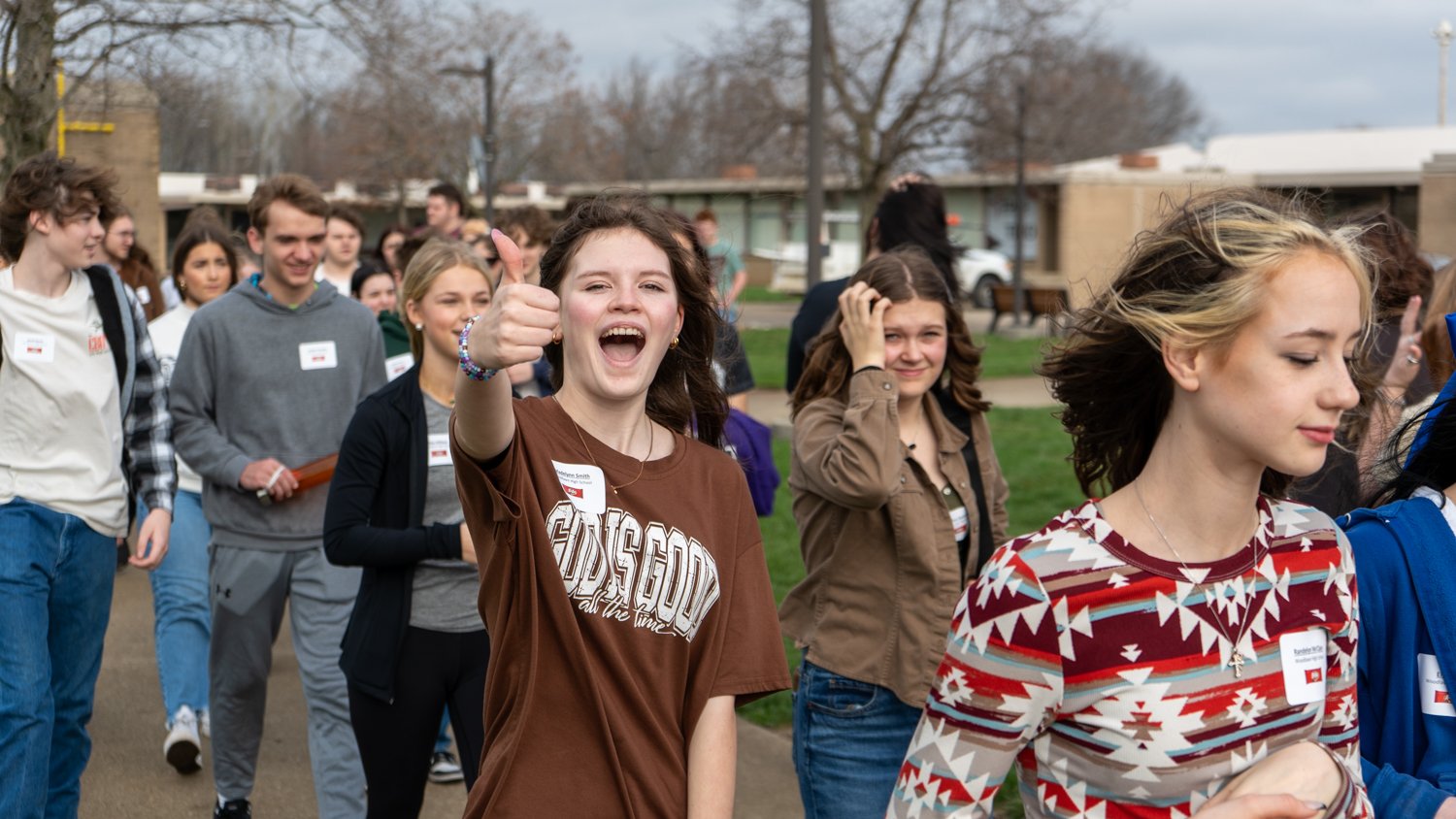 A group of high school students walk together across a college campus during a visit, wearing name tags. In the foreground, one student smiles and gives a thumbs-up toward the camera while others chat and walk behind them on a cloudy day.