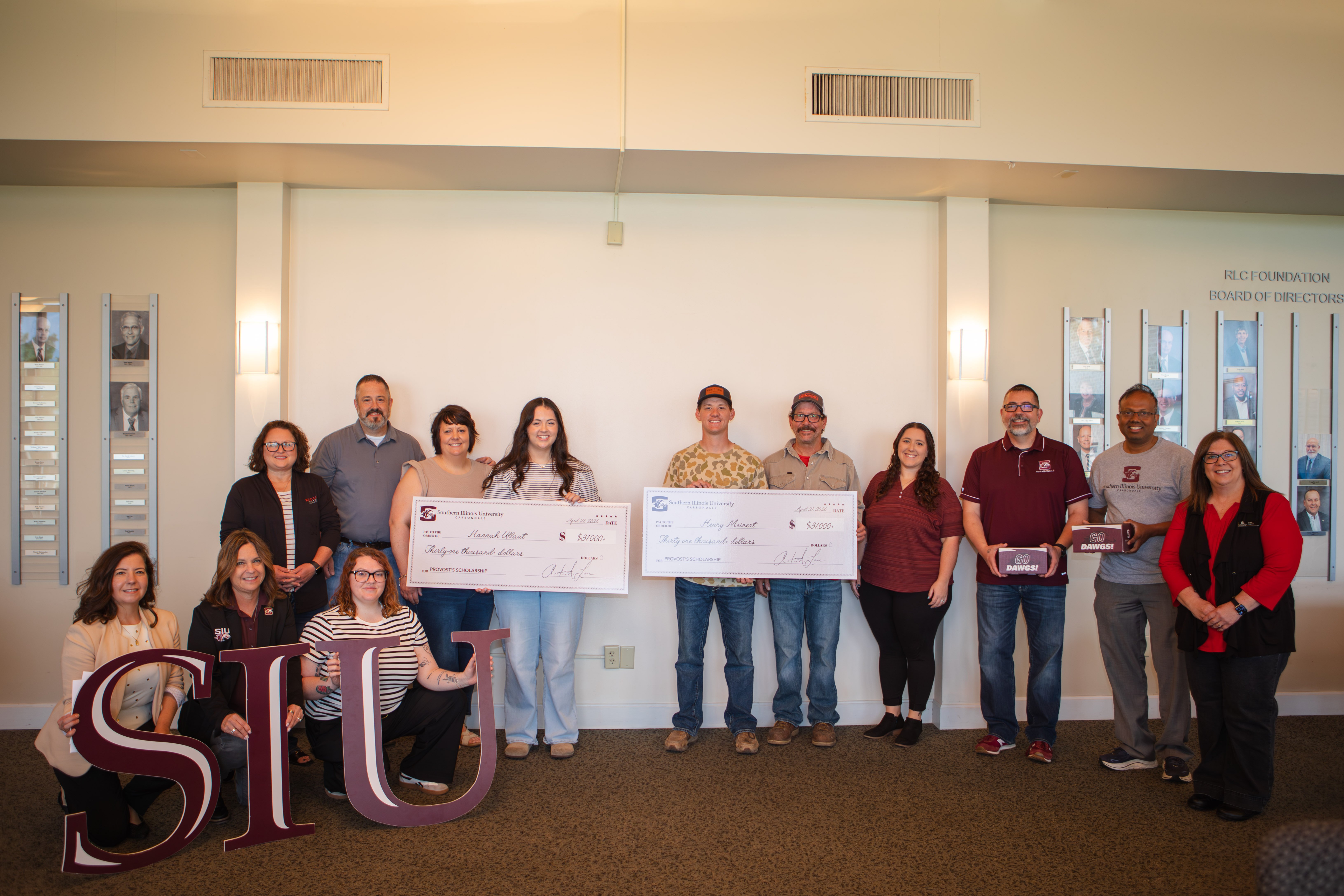 At the center, students Hannah Utlaut and Henry Meinert stand holding large ceremonial checks from Southern Illinois University for the Provost’s Scholarship. They are joined by RLC President Lori Ragland, SIU officials, and their families. On the far left, three women are kneeling behind large, maroon "SIU" letters. Several individuals on the right are holding maroon gift boxes labeled "Go Dawgs!".