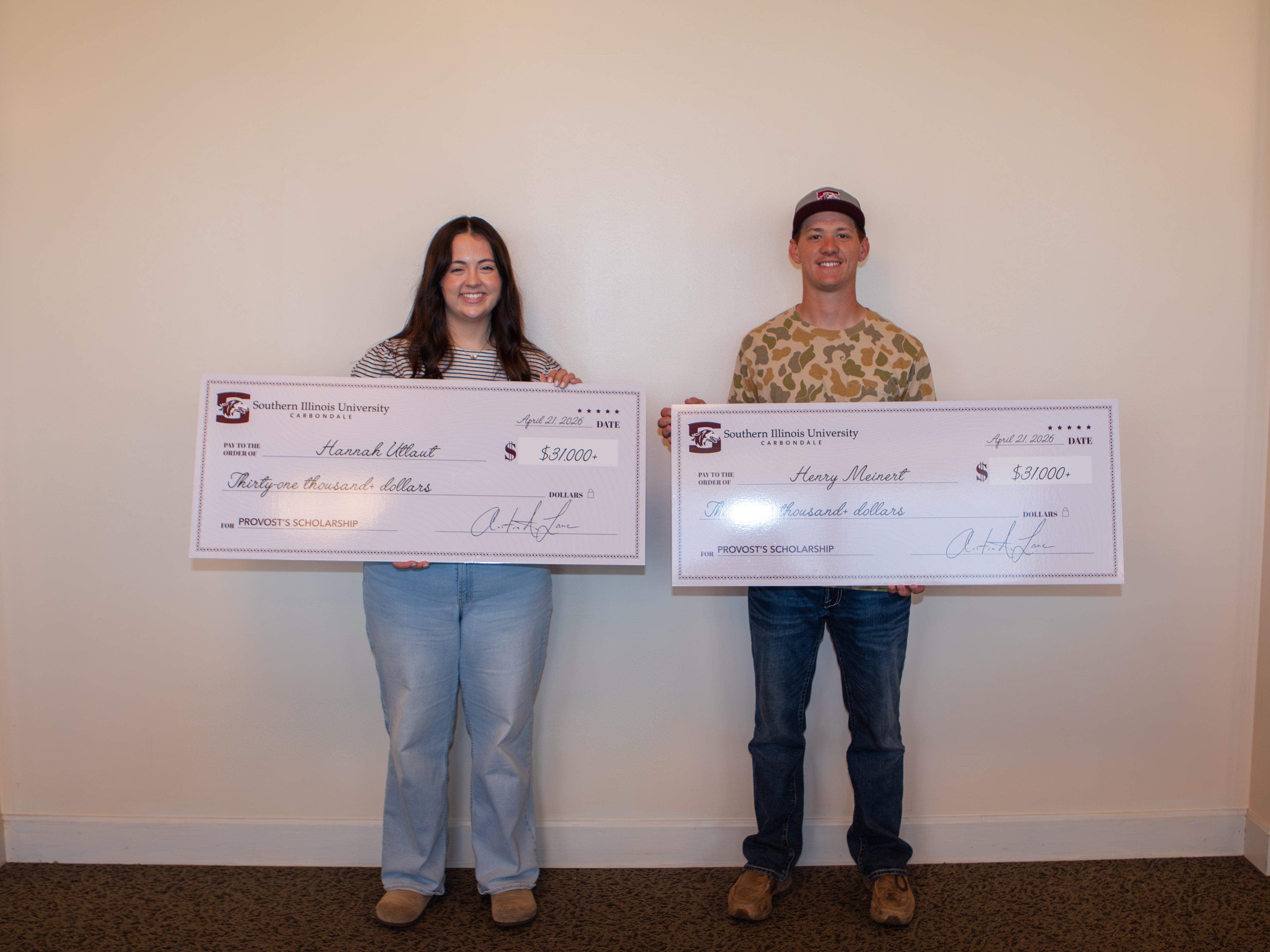 Hannah Utlaut and Henry Meinert stand side-by-side against a plain white wall, each holding a large ceremonial check from Southern Illinois University Carbondale. Both checks are made out for thirty-one thousand dollars for the Provost's Scholarship. Hannah, on the left, is wearing a striped shirt and light-colored jeans, while Henry, on the right, is wearing a camouflage-patterned shirt and a baseball cap.