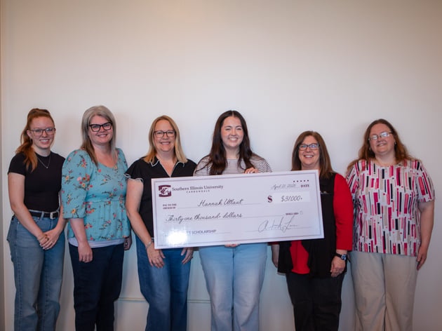 six women stand against a plain white wall holding a large ceremonial check from Southern Illinois University Carbondale for the Provost's Scholarship. Pictured from left to right are Event Coordinator Sydney Clayton, Executive Assistant of Instruction & Student Affairs Karessa Keyser, Executive Director of Administrative Services Cathy DeJarnette, student Hannah Utlaut, President Lori Ragland, and Executive Assistant to the President Mary Huggins. Hannah Utlaut stands in the center, smiling and holding the check, which is made out for thirty-one thousand dollars.