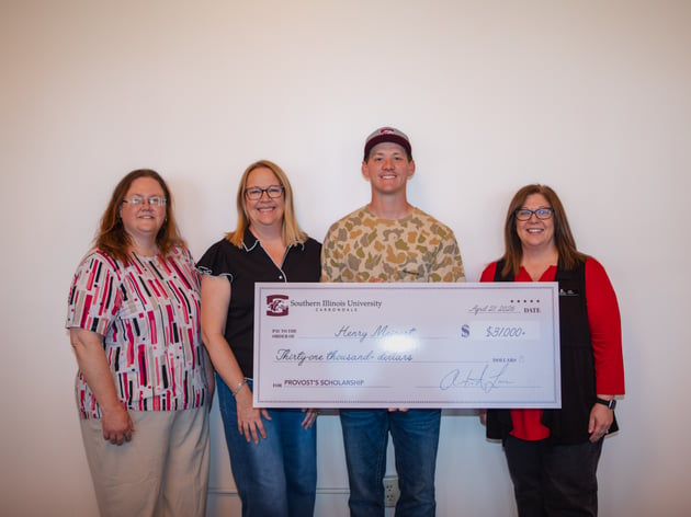 four individuals are pictured standing against a plain white wall while holding a large ceremonial check from Southern Illinois University Carbondale for the Provost's Scholarship. Pictured from left to right are Executive Assistant to the President Mary Huggins, Executive Director of Administrative Services Cathy DeJarnette, student Henry Meinert, and President Lori Ragland. Henry Meinert stands in the center wearing a camouflage shirt and a baseball cap, holding the check which is made out for thirty-one thousand dollars.
