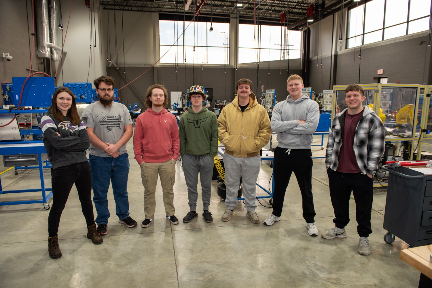 Seven apprentices from the 2026 Industrial Mechatronics and Maintenance cohort stand side-by-side in a modern, high-ceilinged industrial training facility. The group, consisting of one woman and six men, is dressed in casual work attire like hoodies and jeans. They are positioned in the center of the Southern Illinois Manufacturing Academy, surrounded by technical equipment including blue industrial workbenches, electrical control modules, and robotic machinery visible in the background. Natural light streams in from large, high-set windows, illuminating the polished concrete floor.