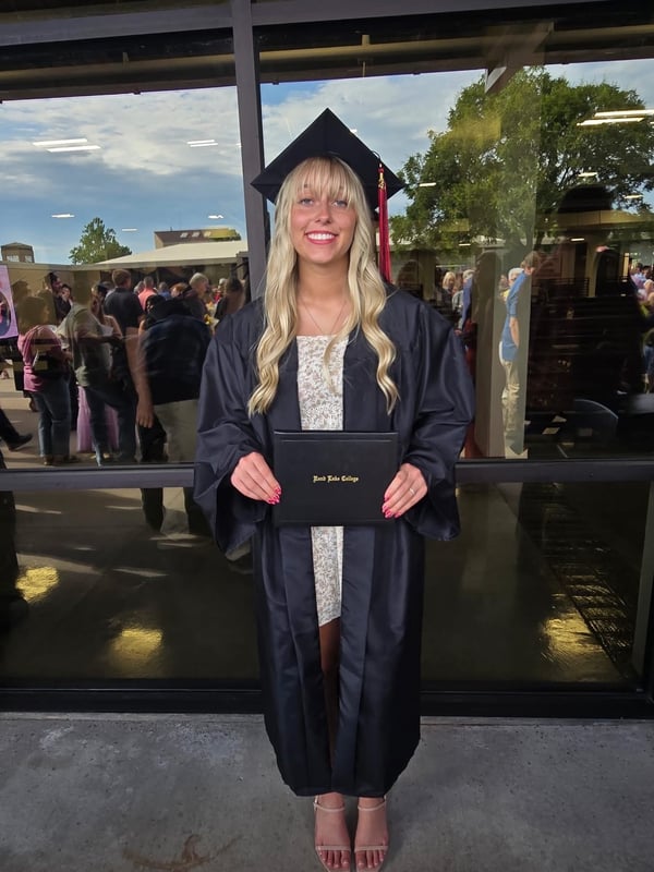 Rend Lake College graduate Bella Morgan smiling in a black graduation cap and gown while holding her diploma cover in front of glass windows on campus.