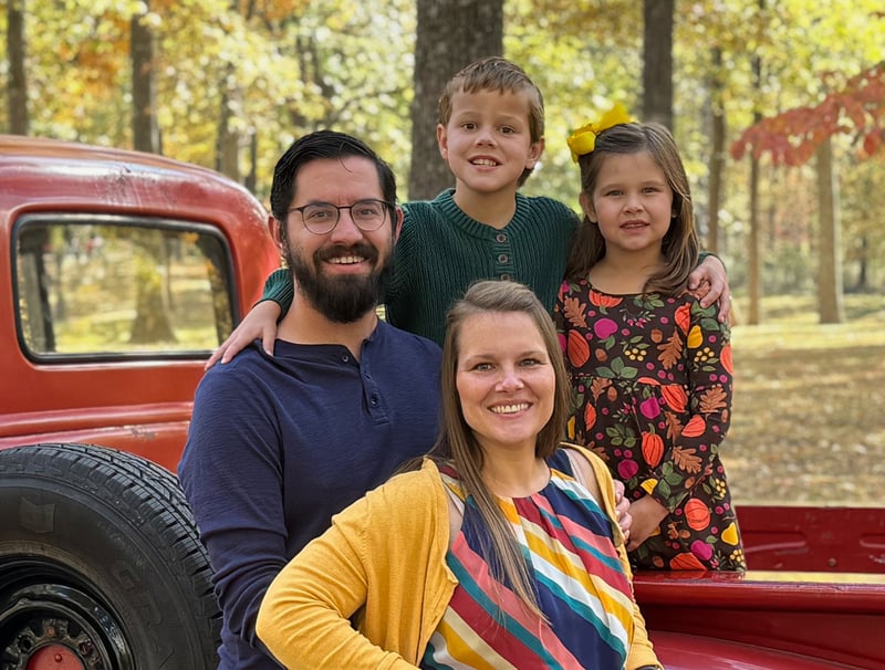 Alt Text: Jonathan McCoy smiling outdoors with his wife and their son and daughter, posing next to a classic red pickup truck with autumn trees in the background.