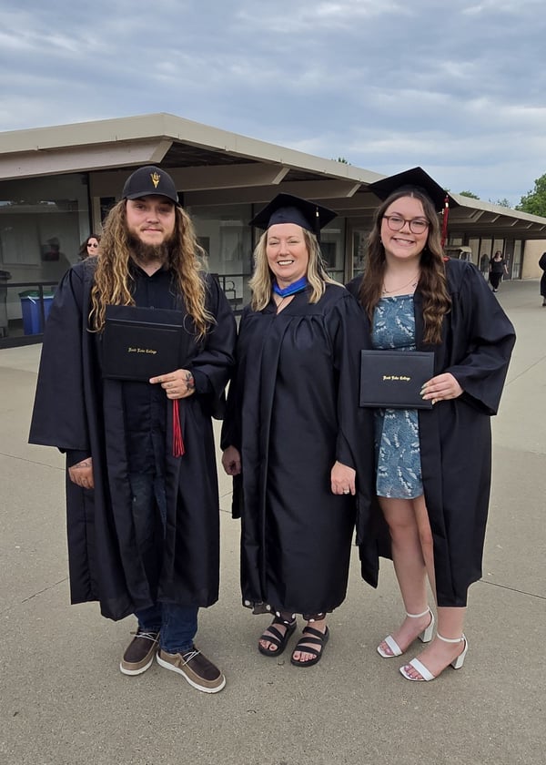 A full-length outdoor photograph showing three people standing side by side in graduation attire on a concrete walkways in front of a campus building. On the far left is Rend Lake College graduate Jonathan Gulley, wearing a black gown, dark jeans, casual shoes, a black baseball cap, and holding a closed diploma cover. In the center is Andrea Banach, Dean of Pinckneyville Campus Advancement and Outreach, wearing a black graduation gown with a blue stole, a mortarboard cap, and black sandals. On the far right is Jonathan's sister, Autumn Gulley, who also graduated that day, wearing a black graduation gown over a dark patterned dress, white heel sandals, a mortarboard cap, and holding a closed diploma cover. All three are smiling toward the camera under an overcast sky.