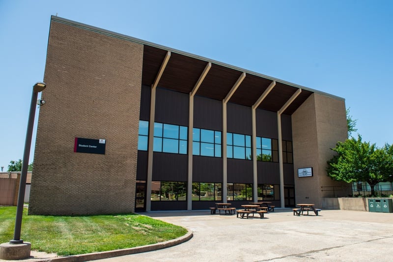 Exterior view of a modern multi-story brick building with large glass windows and an overhanging roof supported by wooden beams, featuring a "Student Center" sign.