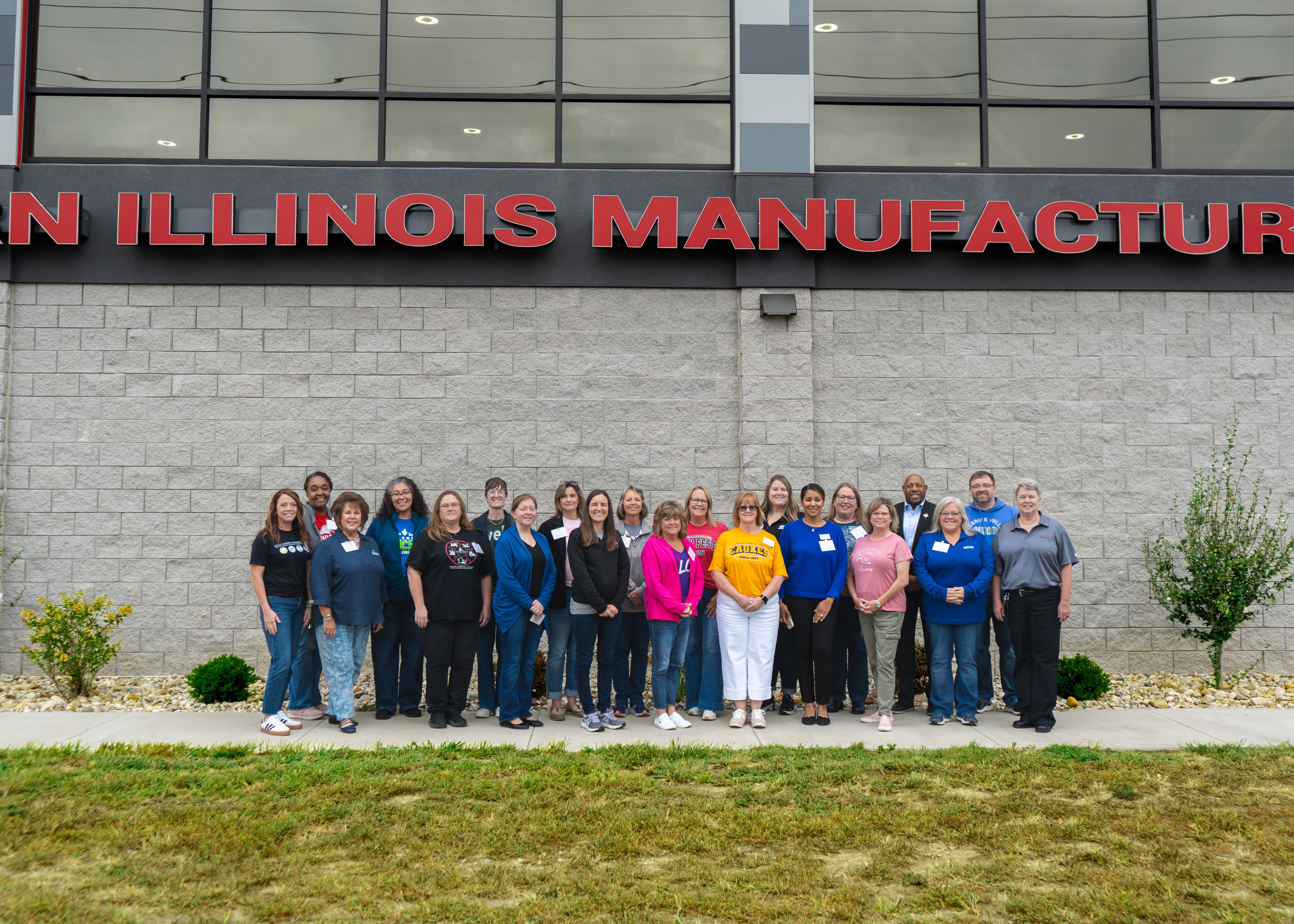 A group of around 20 attendees standing in a line outside a concrete block building with red lettering that reads "SOUTHERN ILLINOIS MANUFACTURING..." on the facade.