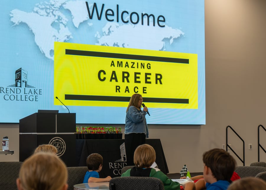 Dr. Lori Ragland, RLC President, speaks into a microphone on stage, welcoming students to the Amazing Career Race in front of a large screen displaying a "Welcome" map graphic and the Rend Lake College logo.