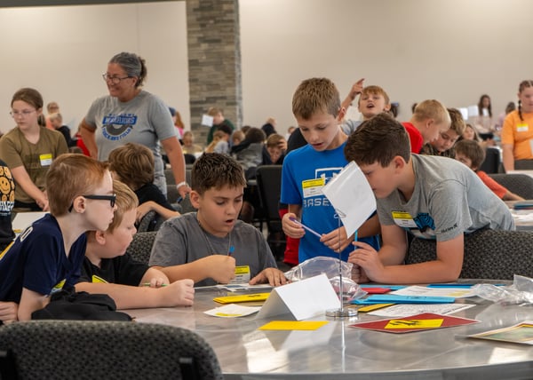 A group of students gathered around a table engaged in a hands-on activity, working with papers, folders, and small props while an adult supervisor looks on in the background.