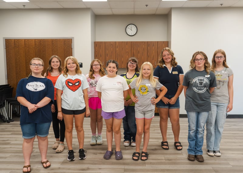 A group of ten young camp participants pose together for a photo in a brightly lit classroom or multipurpose room.