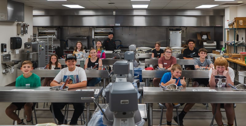 A group of camp participants and a chef instructor pose at stainless steel prep tables in a commercial kitchen training facility.