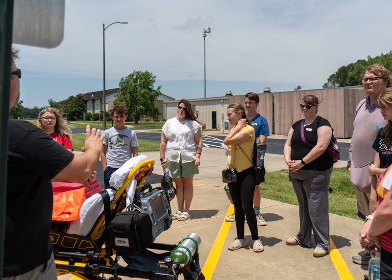 A group of camp participants listens to an instructor give a demonstration next to an ambulance stretcher loaded with medical equipment outdoors.