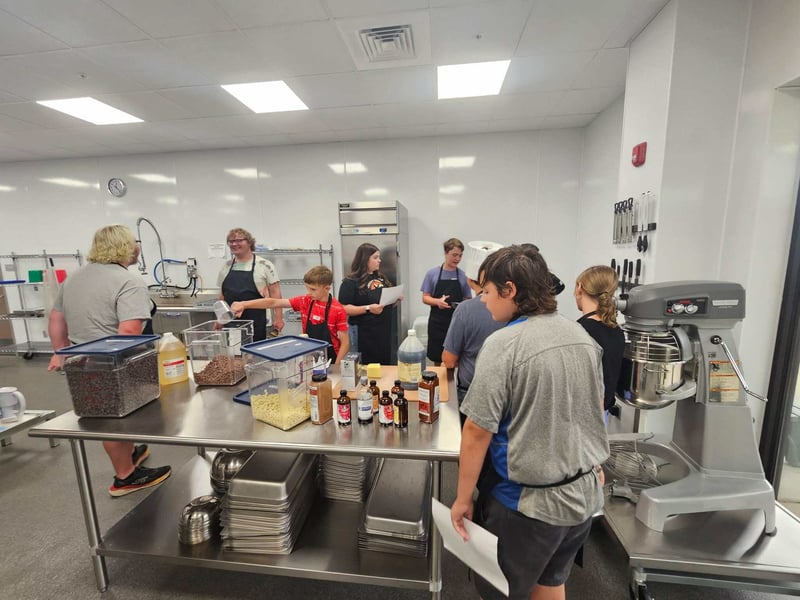 Camp participants gather around a stainless steel prep table with ingredients and baking supplies in a commercial kitchen classroom.