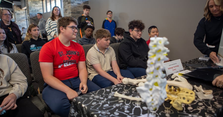 A group of teenage students sits at a career fair, attentively viewing a "Radiologic Technology Careers" exhibit. The table, draped in a skeleton-print cloth, features anatomical models of a human spine and bones. A representative stands behind the display in a modern room with stone accents and large windows.