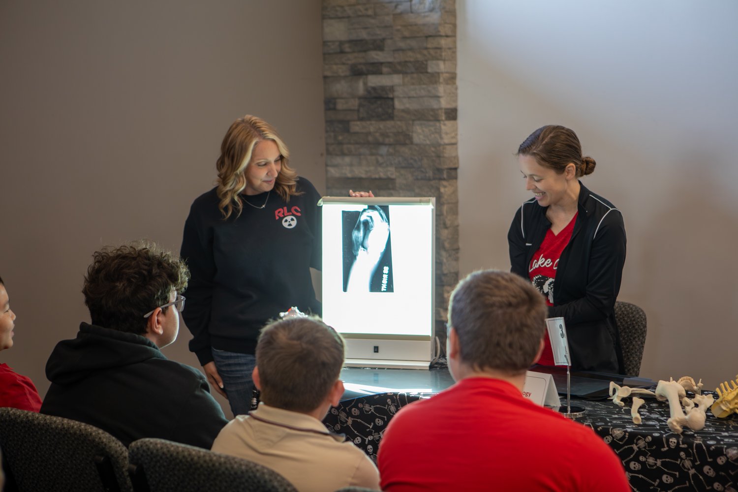 Two instructors demonstrate radiologic technology to seated students by displaying an X-ray on a light box. Anatomical bone models rest on a skeleton-print tablecloth in front of them.