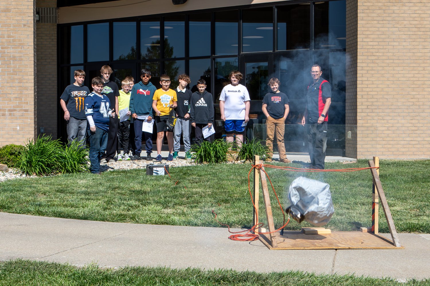 A group of students and an instructor watch an outdoor demonstration of a deployed vehicle airbag. Smoke rises from the airbag assembly, which is secured to a wooden frame on a sidewalk in front of a brick building.