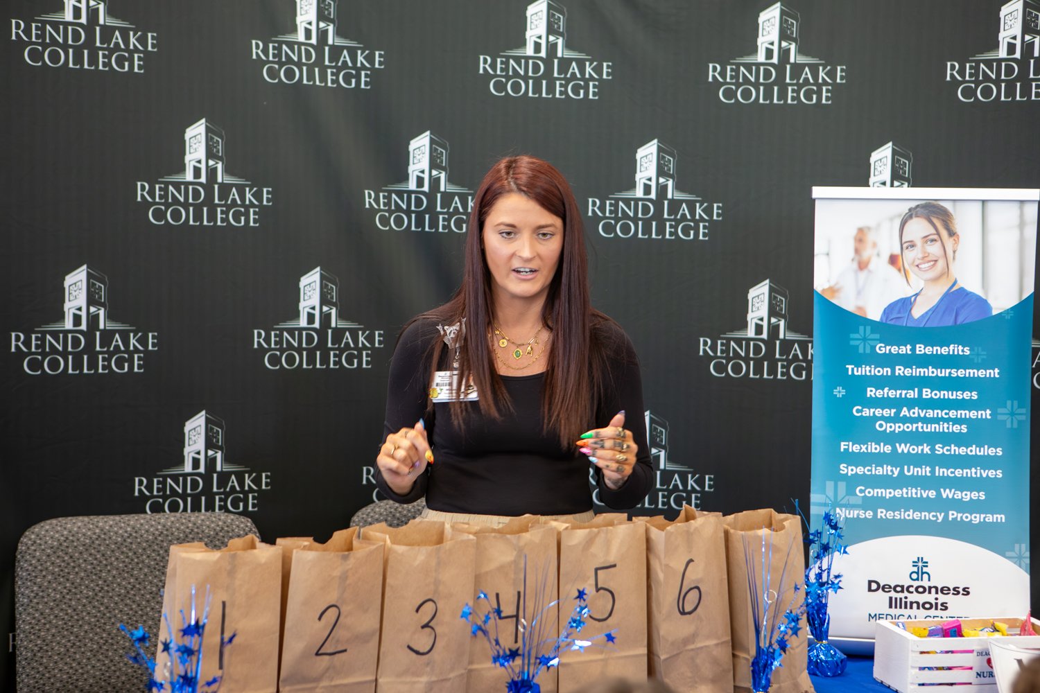  A representative from Deaconess Illinois Medical Center stands in front of a "Rend Lake College" backdrop. She presents a row of brown paper bags numbered one through six, decorated with blue star tinsel, at a career fair table.