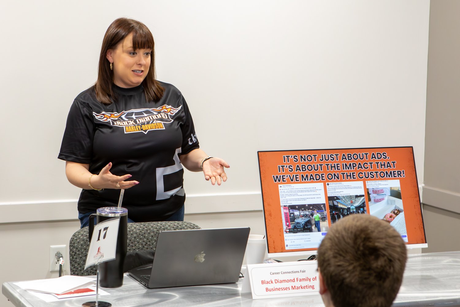 A woman wearing a Black Diamond Harley-Davidson shirt presents to a student at a career fair. She stands behind a table featuring a laptop and an orange display sign that reads, "IT'S NOT JUST ABOUT ADS. IT'S ABOUT THE IMPACT THAT WE'VE MADE ON THE CUSTOMER!" A small sign on the table identifies the booth as "Black Diamond Family of Businesses Marketing."