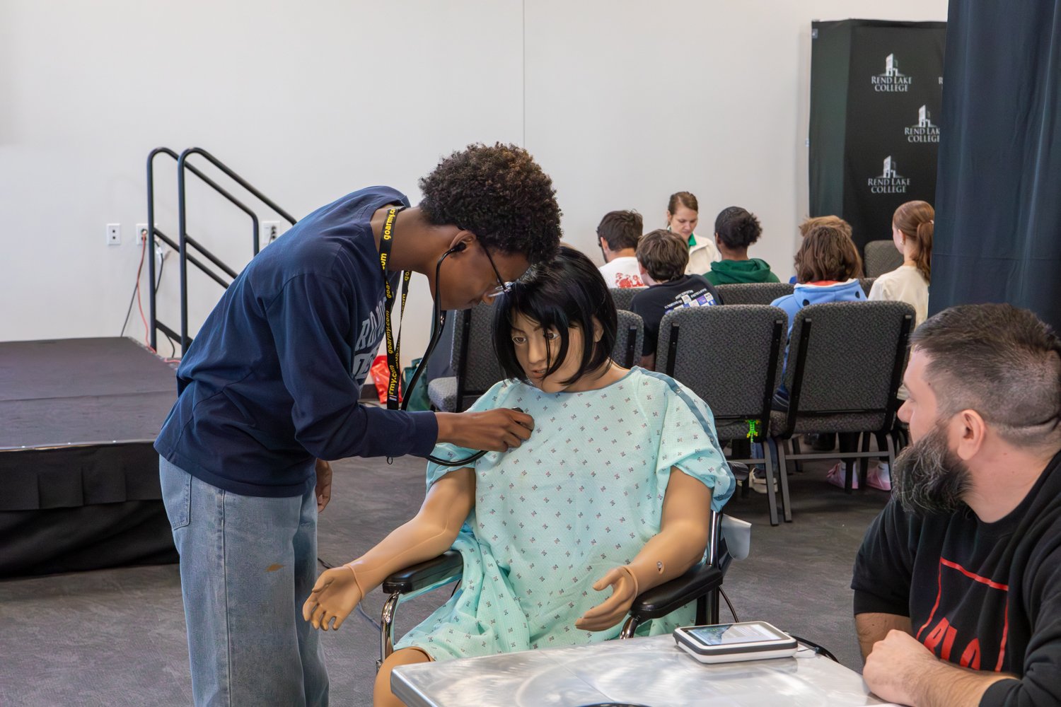  A student uses a stethoscope to listen to the chest of a medical mannequin wearing a hospital gown. A man observes the demonstration from a nearby table, while other students sit in the background of the event space.
