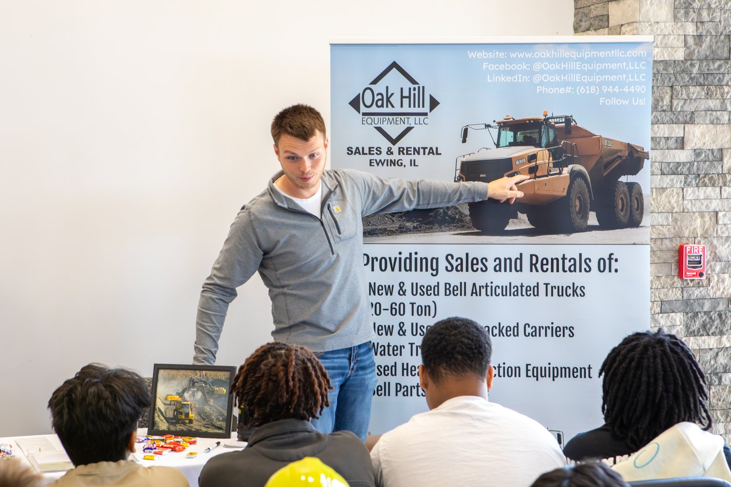 A representative from Oak Hill Equipment, LLC points to a banner showcasing heavy machinery while presenting to a group of students. A framed photo of construction equipment and candy sit on the table in front of him.