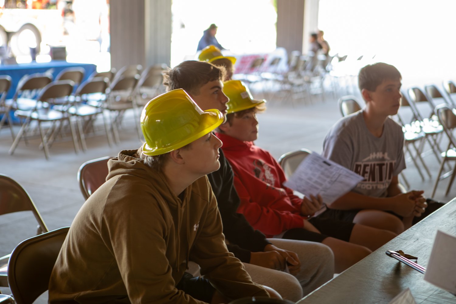 A row of teenage boys sits in folding chairs at an open-air event, focused on a presentation. Several students wear bright yellow hard hats, and one holds an information sheet.