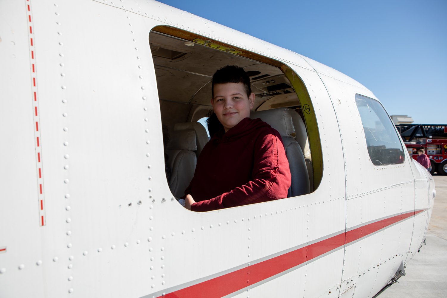  A teenage boy in a maroon hoodie smiles while sitting inside a white and red small aircraft. He is visible through an open cabin door against a clear blue sky.