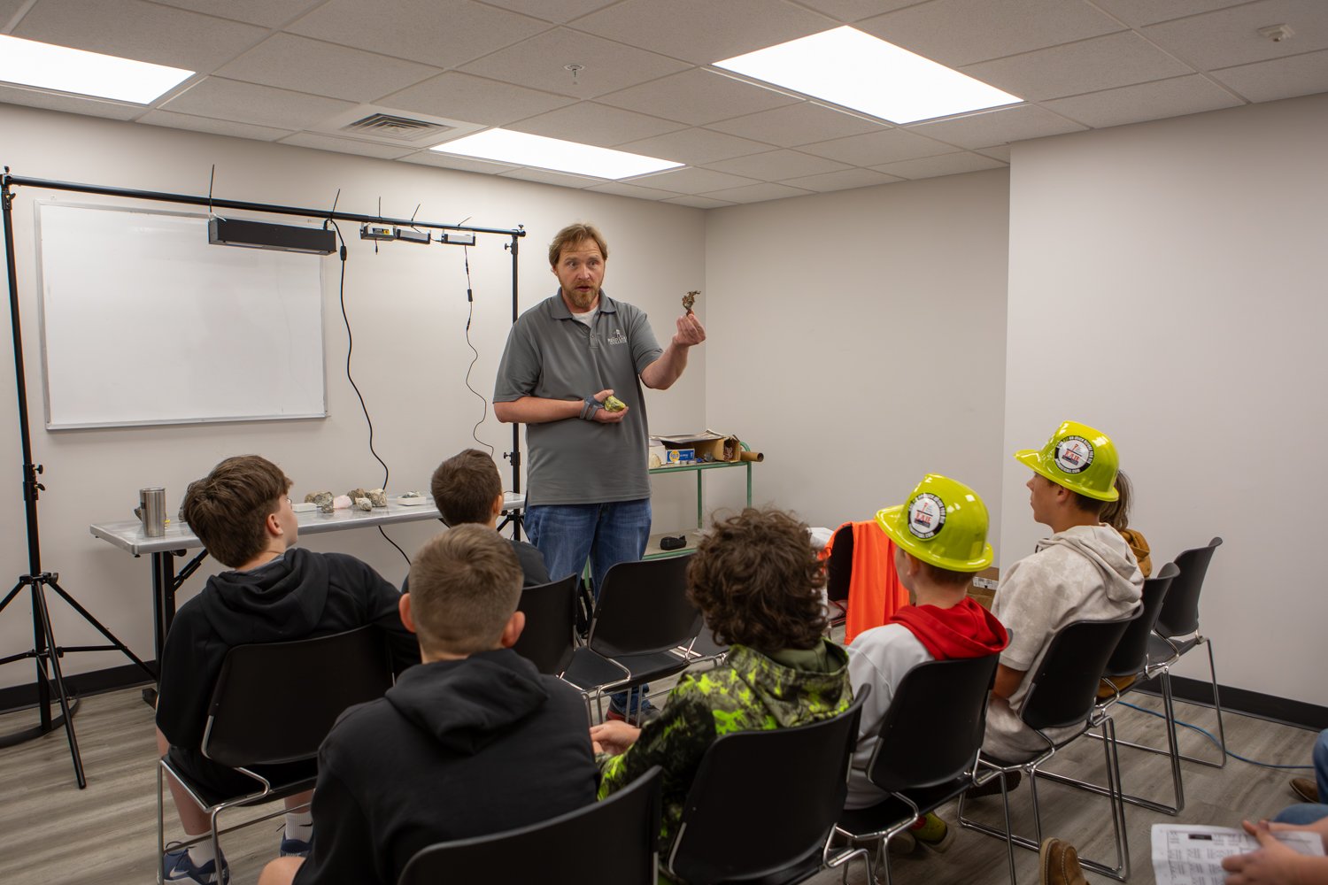 A man in a grey polo shirt shows mineral samples to seated students in a classroom. Two students wear bright yellow hard hats, and a table in the background holds additional geological specimens.
