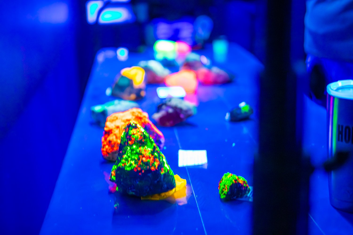 Various fluorescent minerals glow in vibrant neon shades of orange, green, and red under a UV blacklight. The specimens are arranged on a dark table, some accompanied by small white identification labels.