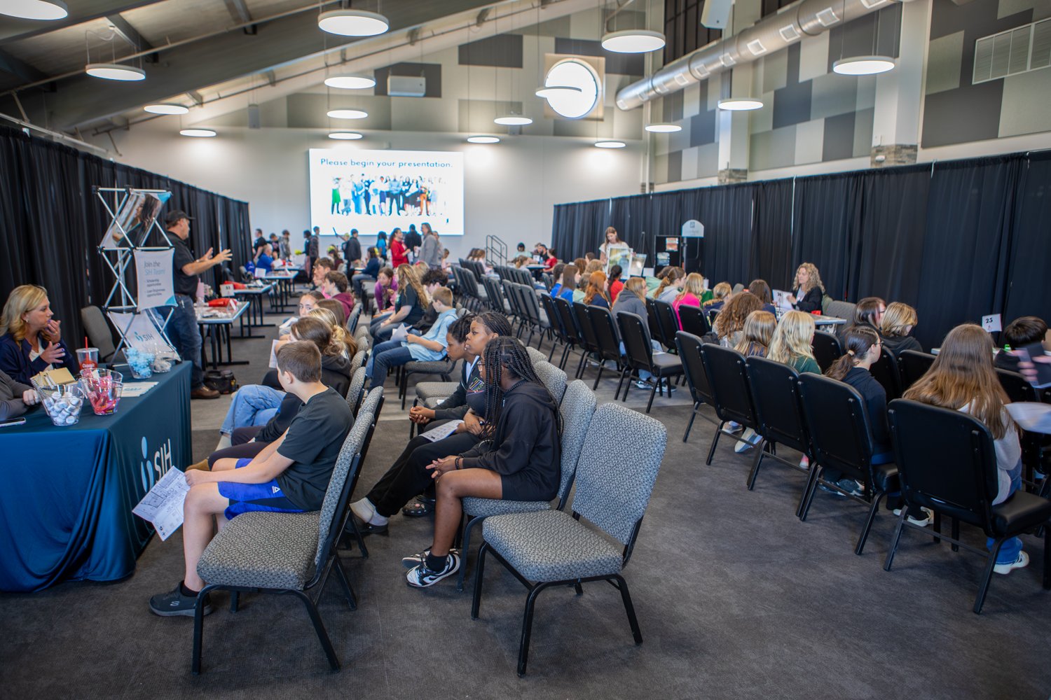 A large convention hall filled with rows of seated students participating in a career event. Organizations, including "SIH," engage with students at booths lined with black curtains, while a large screen at the front reads, "Please begin your presentation."