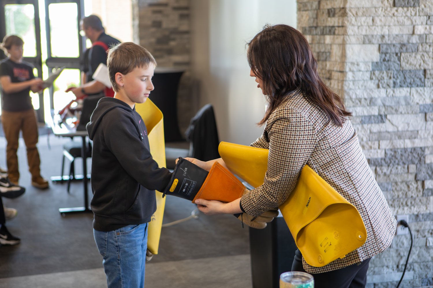 A woman helps a young boy try on heavy-duty electrical safety gloves and yellow insulated sleeves at a career event. They stand in a bright room next to a stone pillar.