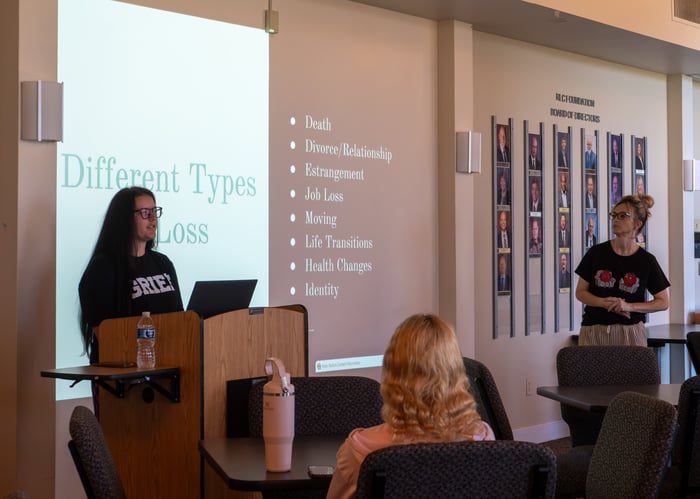 Two presenters stand at the front of a lecture hall in front of a projection slide listing "Different Types of Loss," including death, divorce, job loss, and life transitions, while students listen from seated tables.