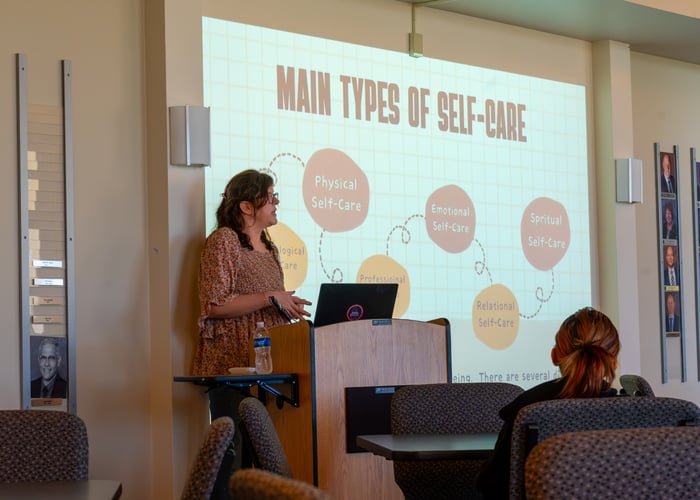 Becca Chavez, a Licensed Professional Counselor, presents at a podium in front of a projection screen displaying a flowchart titled "Main Types of Self-Care" with circles for physical, emotional, spiritual, and relational self-care.