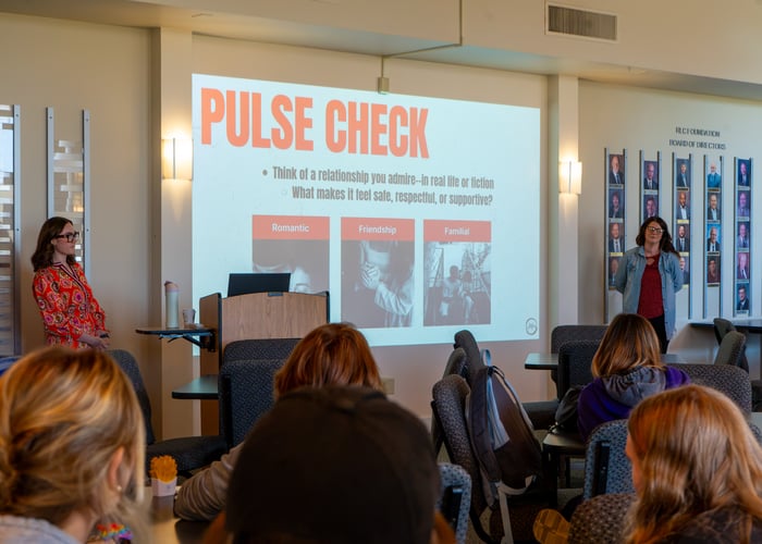 Molly Donoho and Regina McKinney from SAFE present to a room of seated students, with a projection screen behind them showing a "Pulse Check" slide featuring categories for romantic, friendship, and familial relationships.