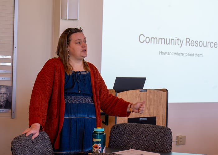 Sarah Draper, Coordinator of Student Success and Wellness, stands at the front of a presentation room speaking to students, with a projection screen behind her reading "Community Resources: How and where to find them!"