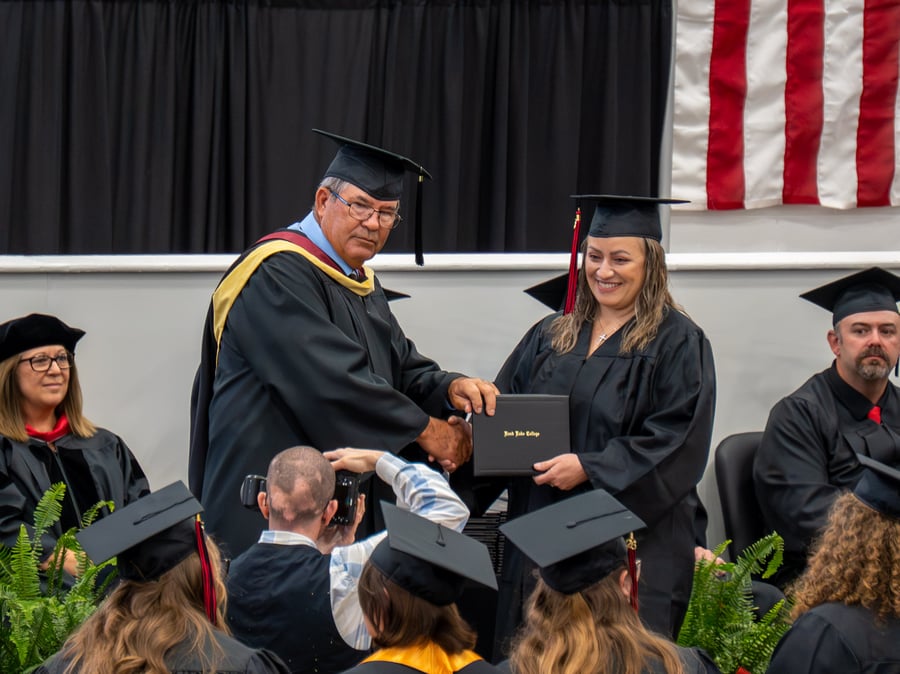 A graduation stage photo capturing a female graduate smiling as she shakes hands with a male college official and receives her diploma cover. Both are wearing black academic regalia and caps. The diploma cover is black with "Rend Lake College" printed in gold script. In the foreground, the backs of other graduates' heads and a photographer taking a picture are visible. In the background, other faculty or stage party members sit in chairs before a black curtain and a section of an American flag.