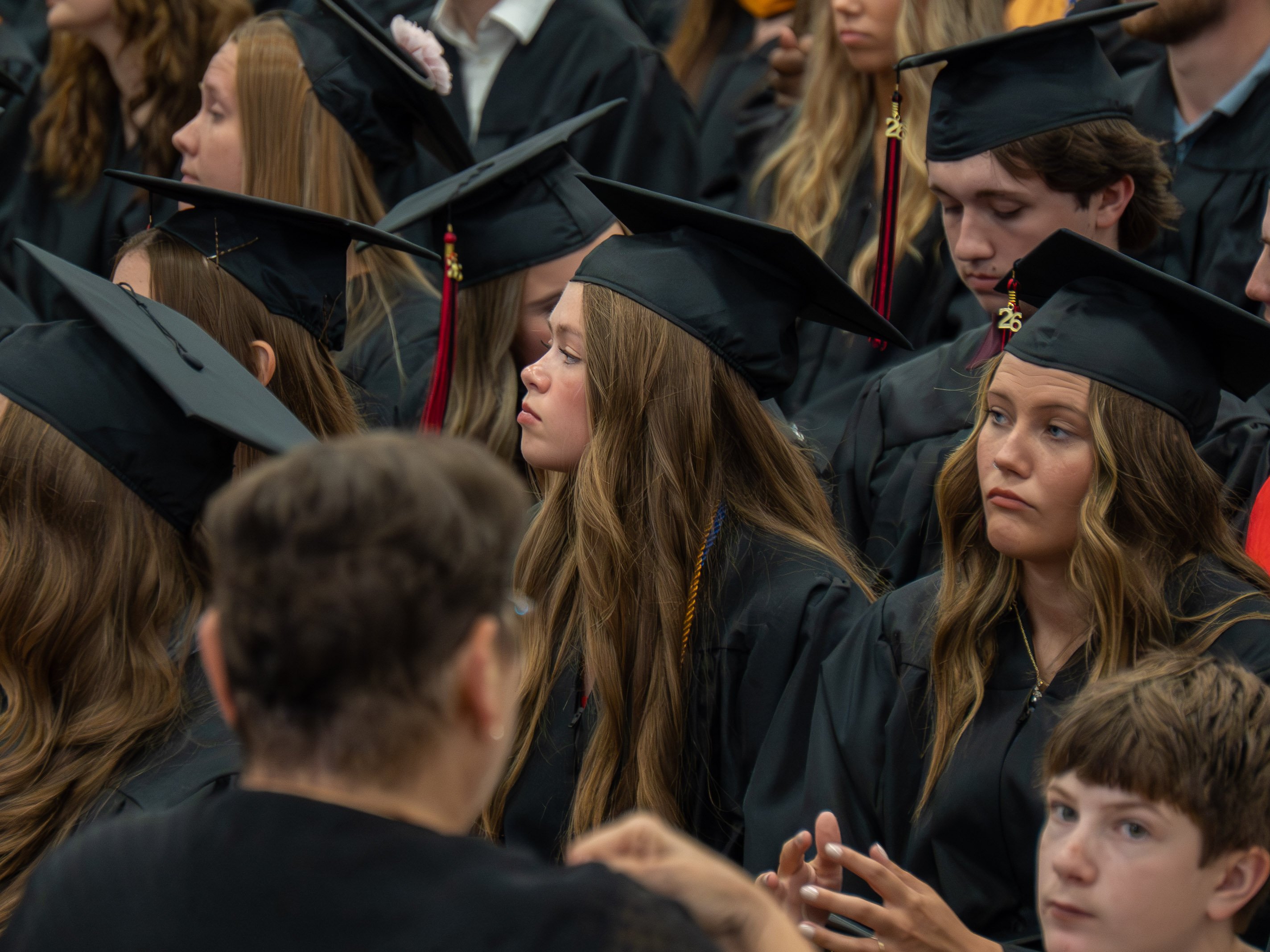A close-up view of a crowded group of graduates seated during a commencement ceremony, all wearing black graduation caps and gowns. In the center, two female graduates with long brown hair look forward attentively, with one making a slight pout or concentrated expression. The surrounding graduates fill the frame, creating a dense pattern of black mortarboards and tassels. The back of an audience member's head is visible in the lower-left foreground.