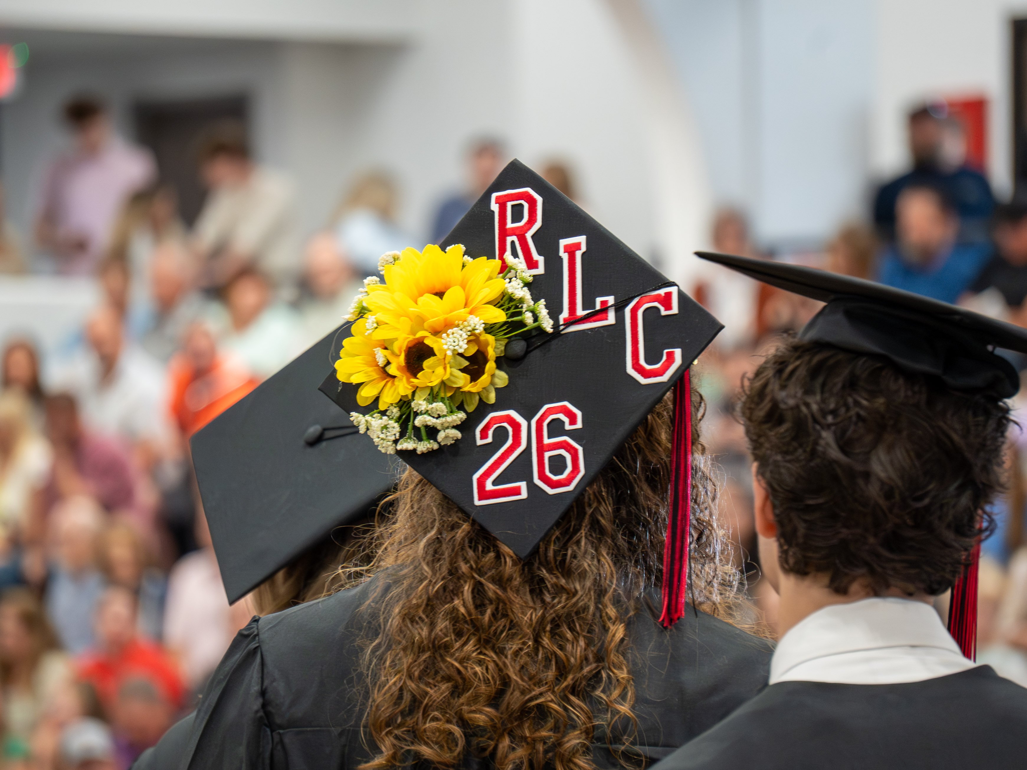 An over-the-shoulder view of a graduate with long, curly brown hair wearing a black graduation cap and gown. The top of the cap is decorated with a cluster of bright yellow sunflowers and white baby's breath flowers, alongside red and white varsity-style letter patches spelling "RLC" and the number "26." To the right, the back of another graduate's head is visible, and the background shows a blurred audience sitting in gym bleachers.