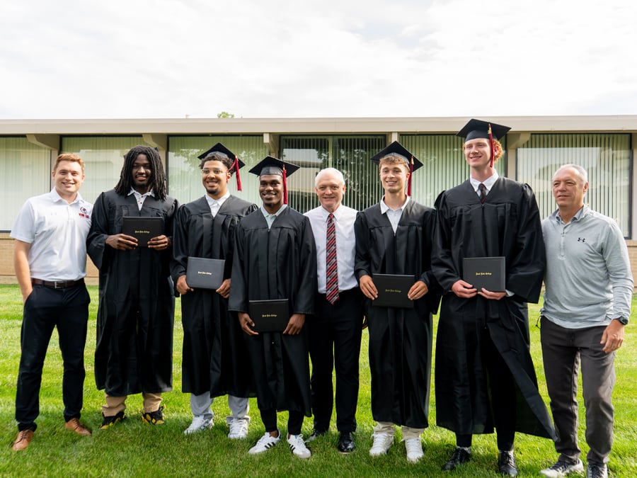 A group photo outdoors on a grassy lawn featuring five male basketball players in graduation caps and gowns holding their diplomas, flanked by their coaches and a teammate.