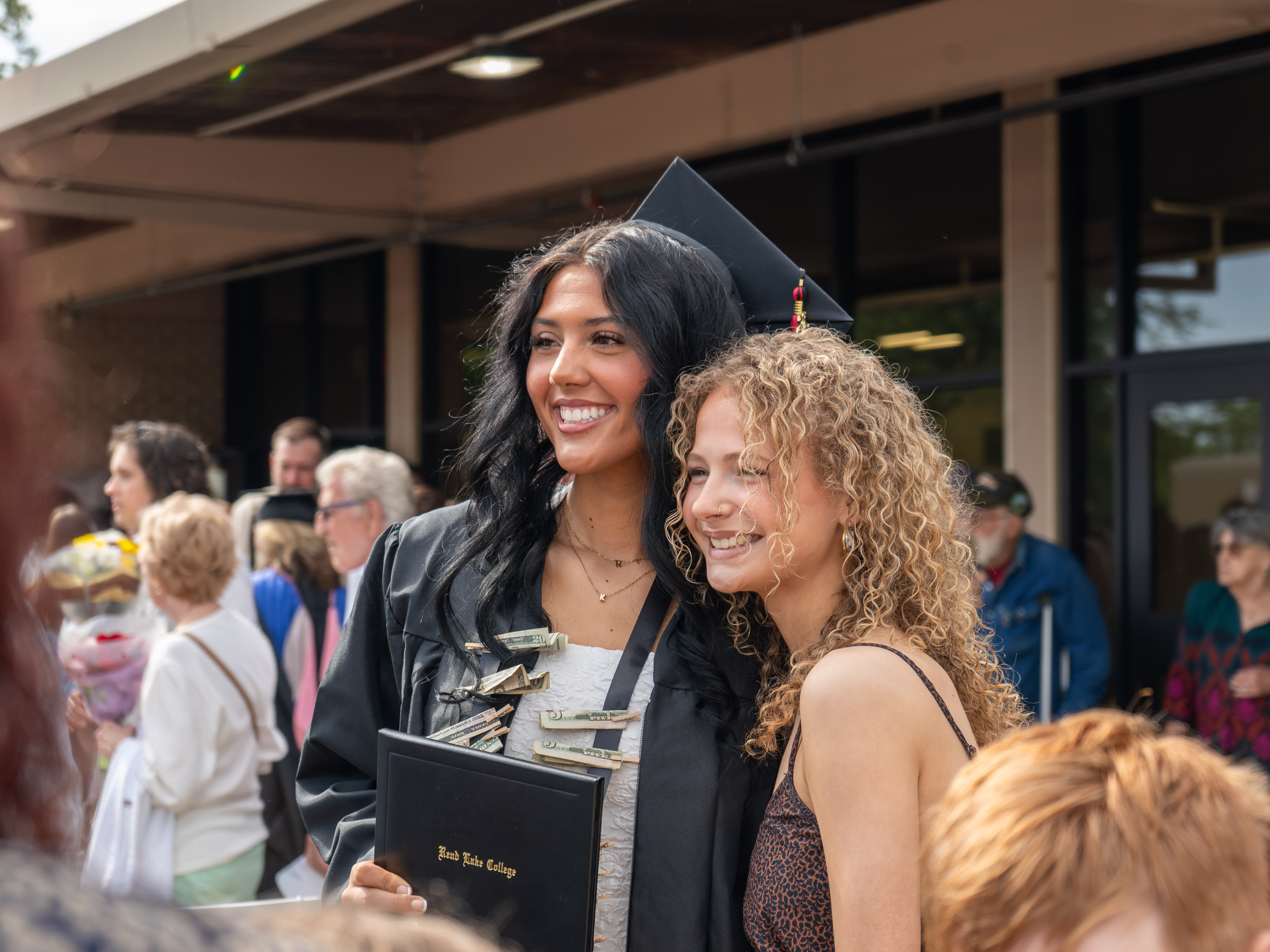 A close-up outdoor photo of a female graduate with long black hair smiling alongside a friend or family member with curly blonde hair. The graduate is wearing a black graduation cap and gown over a white dress, holding a black Rend Lake College diploma cover, and wearing a black ribbon lei pinned with rolled-up dollar bills. Both are smiling brightly and looking slightly to the side. In the background, a crowded campus walkway is filled with other ceremony attendees under the awning of a building.