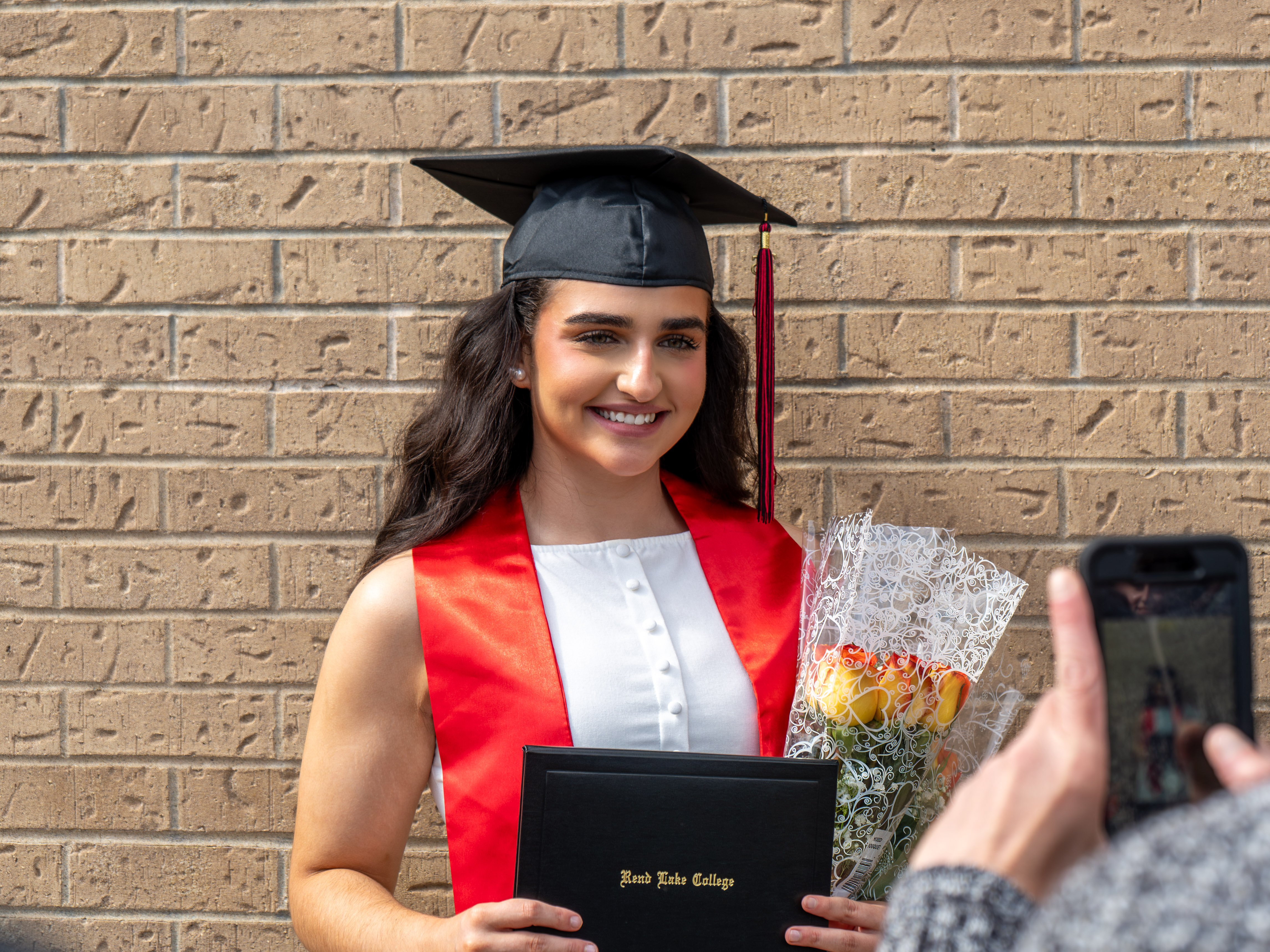 A female graduate smiling as someone takes her photo with a smartphone in the foreground. She is standing outdoors in front of a textured tan brick wall, wearing a black graduation cap, a white top, and a bright red graduation stole. She holds a black Rend Lake College diploma cover in both hands along with a wrapped bouquet of yellow and orange roses.