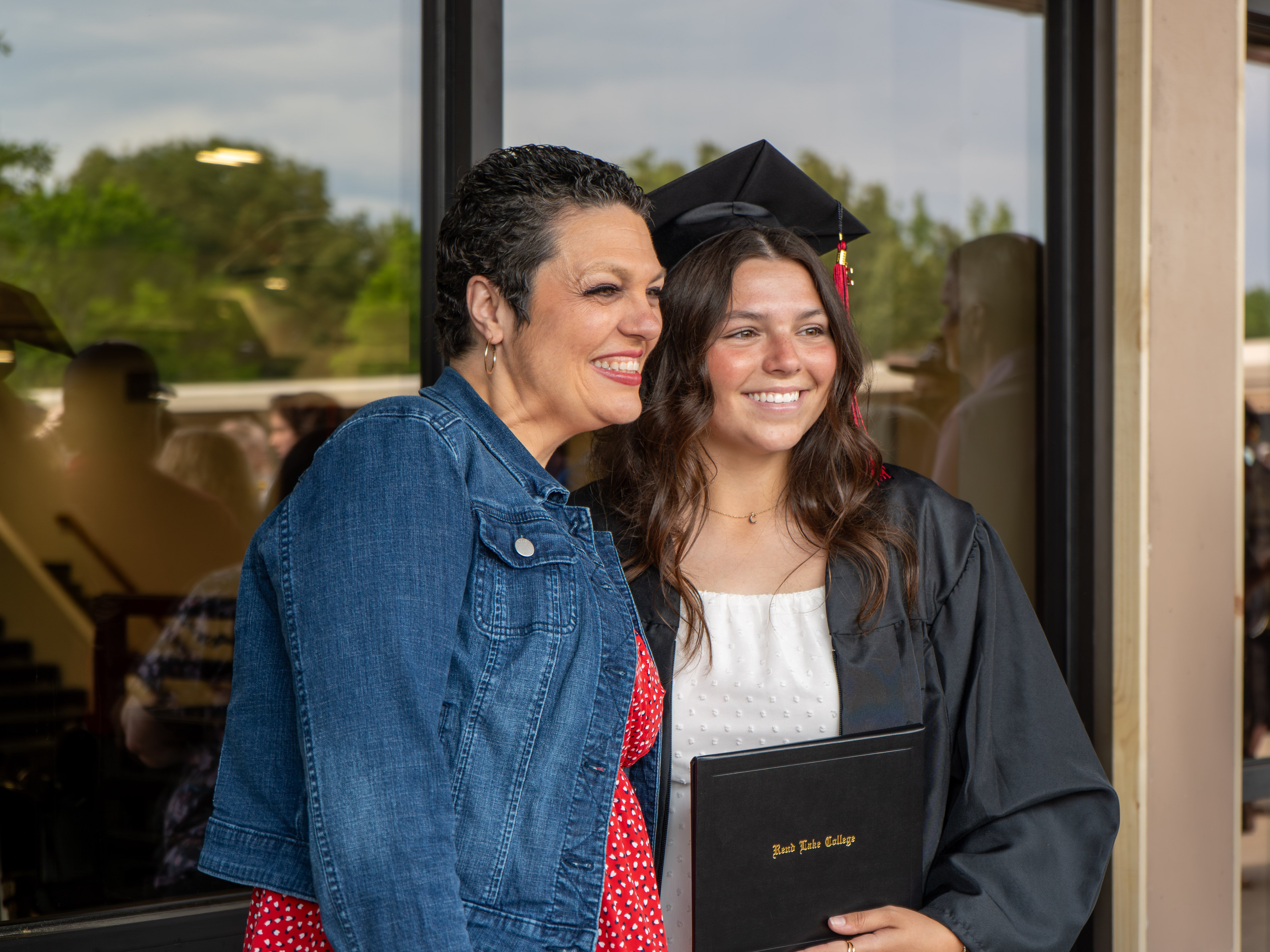 An outdoor close-up photo of a female graduate smiling brightly alongside an older woman after a commencement ceremony. The graduate wears a black graduation cap and gown over a white patterned top and holds a black Rend Lake College diploma cover in her hand. The woman next to her has short dark hair and smiles warmly while wearing a blue denim jacket over a red and white polka-dot dress. They are standing in front of reflective glass windows that capture the faint reflection of other attendees outdoors.