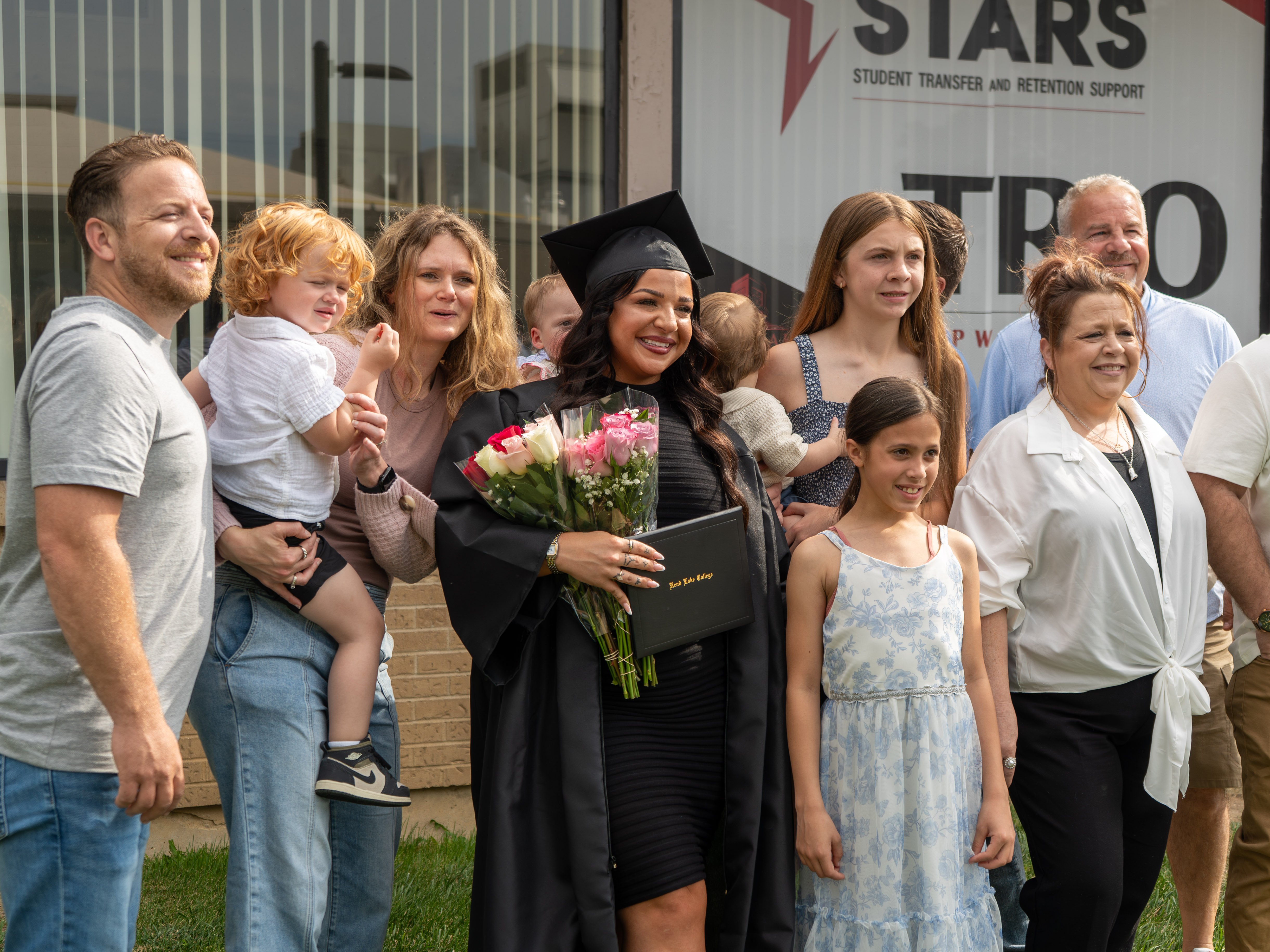 A large group photo outdoors on a campus lawn featuring a female graduate smiling brightly and posing with her family after a commencement ceremony. The graduate stands in the center wearing a black graduation cap and gown, holding a black Rend Lake College diploma cover and a bouquet of pink and white roses. She is surrounded by multiple adults and children, including a toddler being held on the left and a young girl in a blue and white patterned dress standing in the front right.