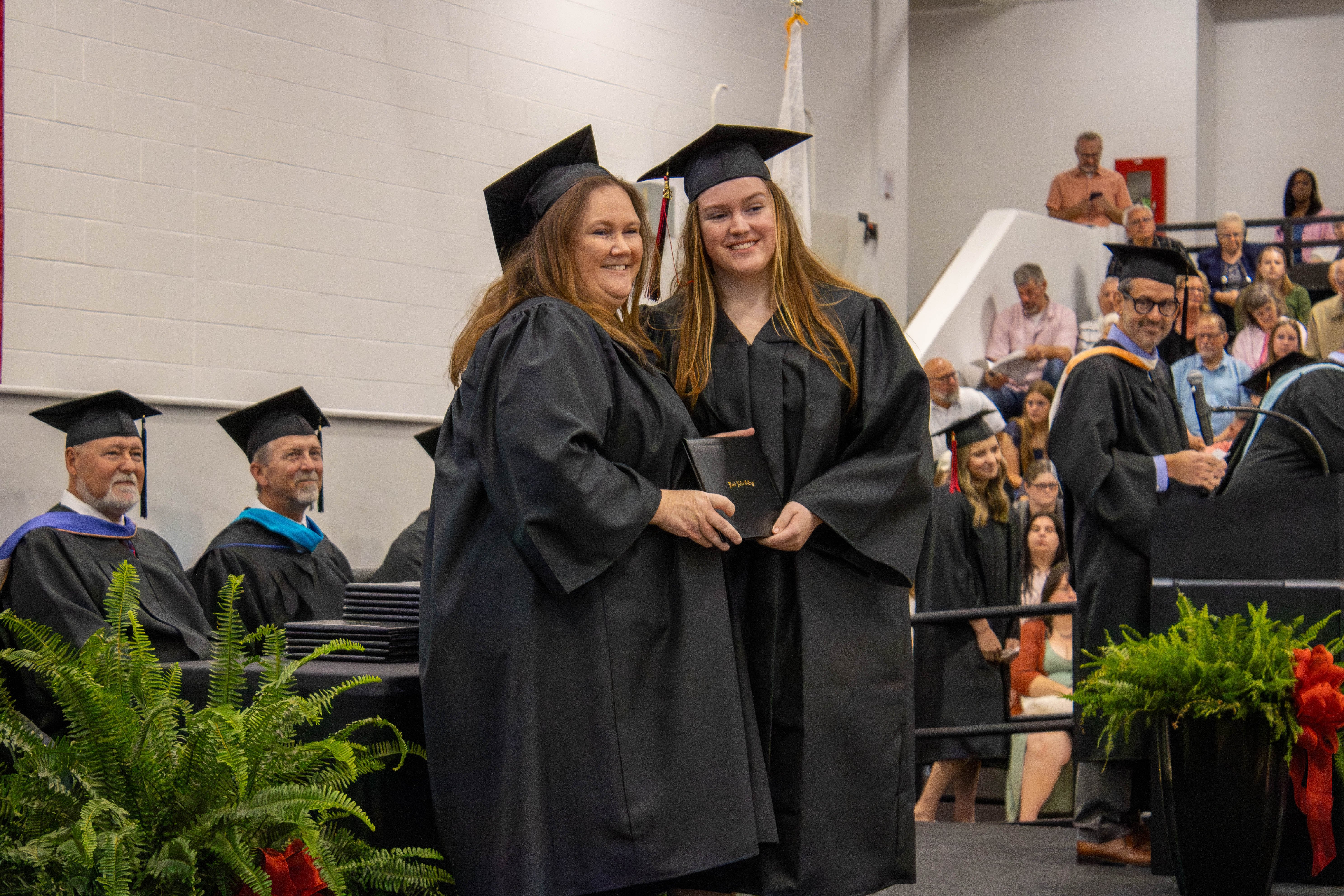 A graduation stage photo capturing two female graduates smiling as they stand side-by-side and hold a black diploma cover together. Both are wearing black graduation gowns and caps. On the left side of the stage, two male faculty members in academic regalia sit behind a table adorned with green ferns. On the right, a faculty member stands at a podium, with a crowded audience seated in gym bleachers visible in the background.