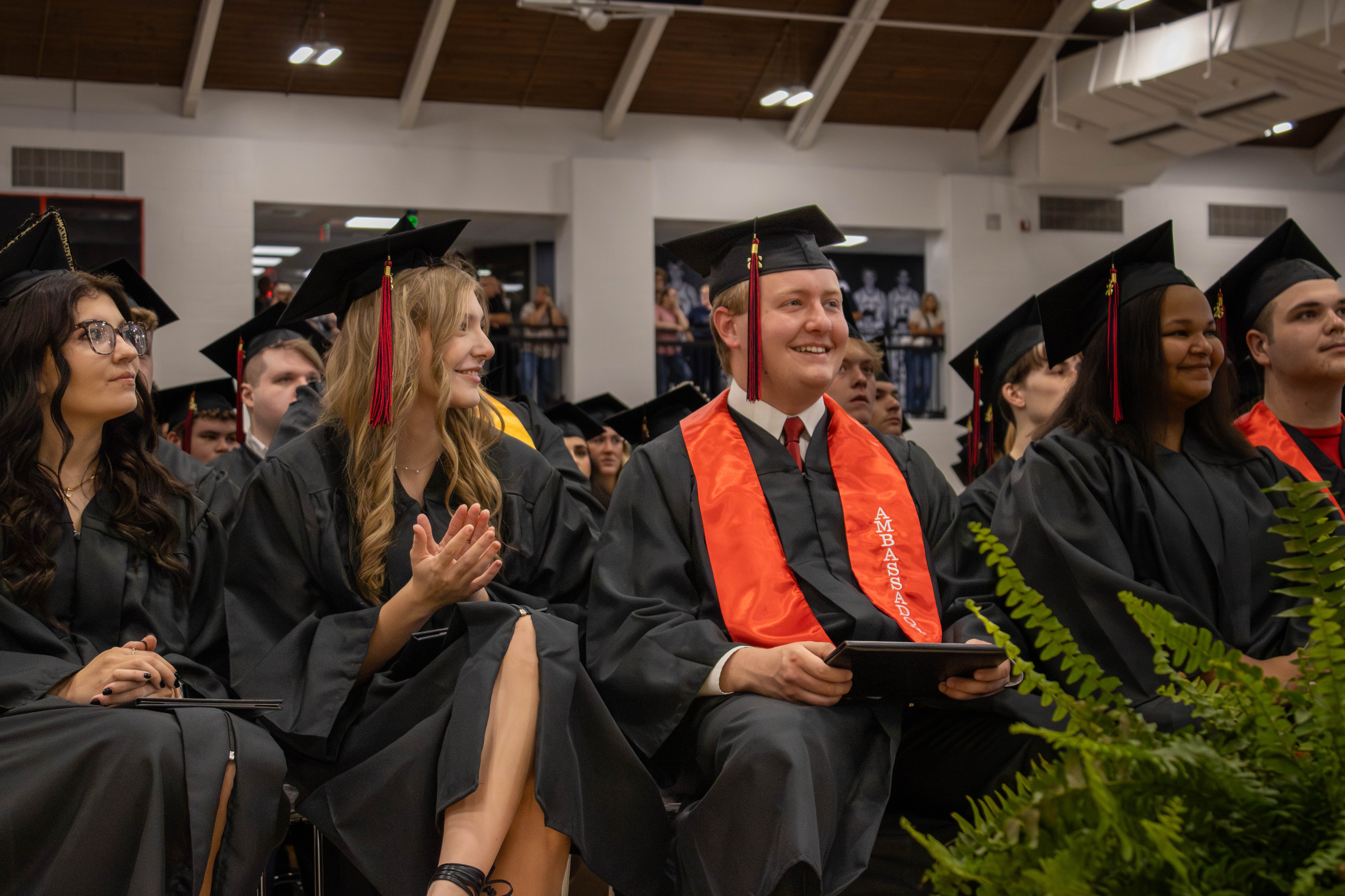 A row of graduates wearing black caps and gowns sitting during a commencement ceremony inside a gymnasium or auditorium. In the center, a male graduate smiles brightly while wearing a vibrant red graduation stole and holding his diploma cover. To his left, a female graduate with blonde hair smiles and claps her hands. Green ferns are visible in the lower right foreground, and other seated graduates can be seen in the background under an exposed beam ceiling.