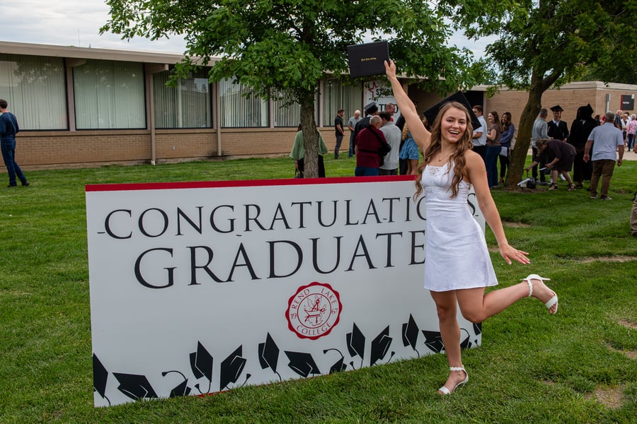 Alt text:  A joyful female graduate poses outside on the campus lawn next to a large white sign that reads "CONGRATULATIONS GRADUATES" in black text, featuring the red Rend Lake College seal and a border of graduation caps. She is wearing a short white dress, white heels, and a black graduation cap, smiling brightly while holding her black diploma cover high in her right hand and playfully kicking one leg back. In the background, other graduates and families gather near a campus building.