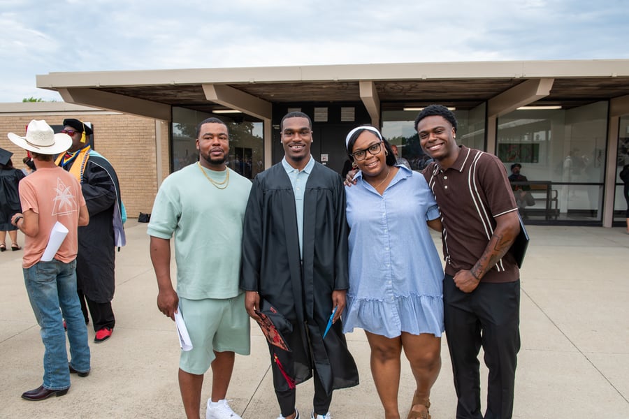 A group of four people smiling together outdoors on a concrete plaza following a commencement ceremony. In the center, a male graduate wears a black graduation gown over a light blue polo shirt, holding his decorated cap in his hand.