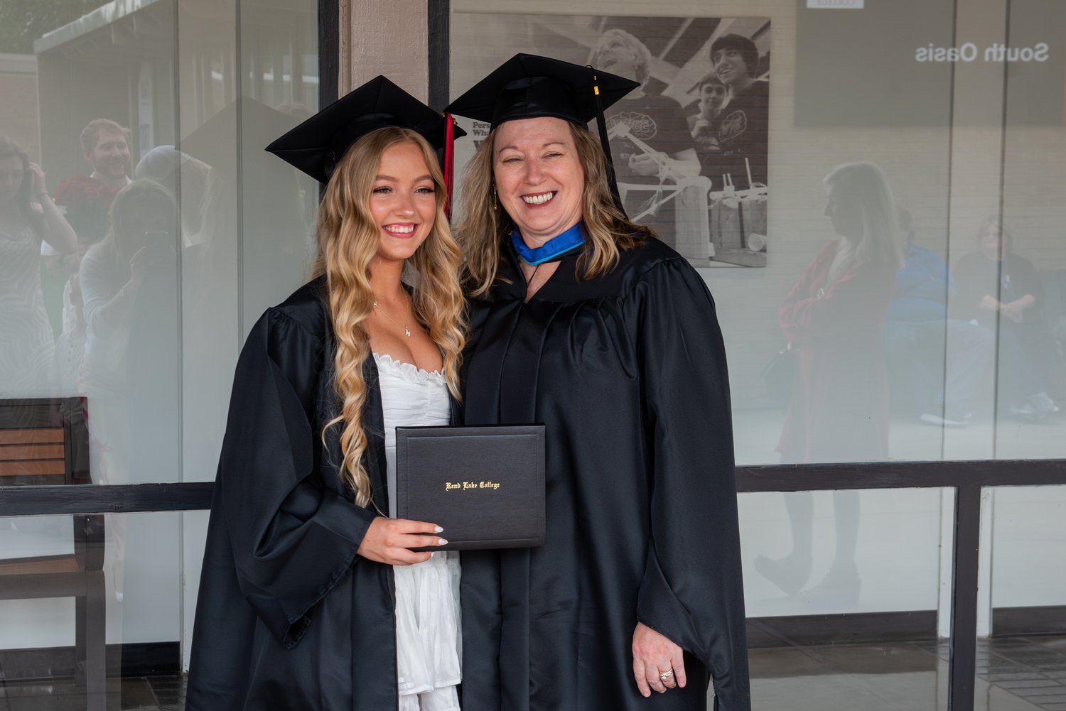 A female graduate and a female faculty member standing close together and smiling outside after a commencement ceremony. The graduate, on the left, has long wavy blonde hair, wears a white dress under an open black graduation gown, and wears a black cap. They are both holding a black Rend Lake College diploma cover together. The faculty member wears a black master's or doctoral gown with a blue hood accent and a black cap, smiling broadly.
