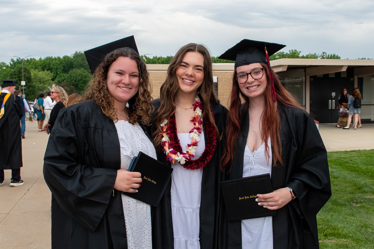 Three female graduates standing close together and smiling on a campus walkway after a commencement ceremony. All three are wearing white dresses underneath open black graduation gowns. The graduate on the left has curly hair and holds a black diploma cover, the graduate in the center wears a maroon and white floral lei around her neck, and the graduate on the right wears glasses and a black graduation cap while holding her diploma cover. In the blurred background, other attendees walk near campus buildings under an overcast sky.
